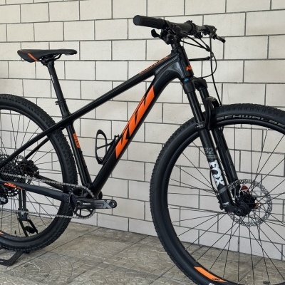 Bicicleta de montanha preta com detalhes laranja encostada em parede de tijolos brancos