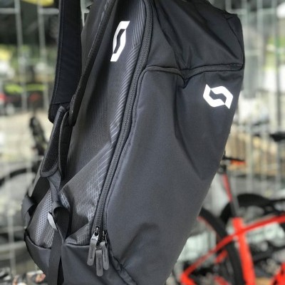 Mochila preta SCOTT com logotipos brancos pendurada em gancho com bicicletas ao fundo