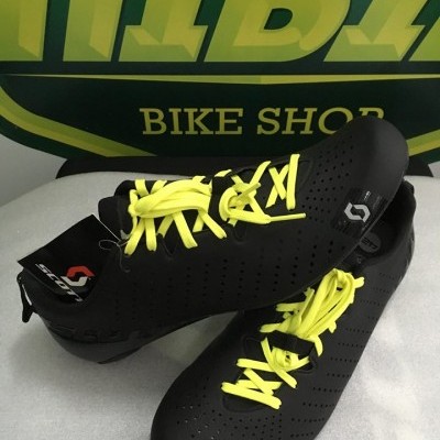 Sapatos desportivos pretos com atacadores amarelos e logotipo Scott em frente a painel verde com texto amarelo