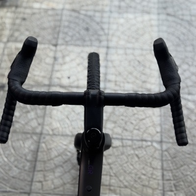Guiador de bicicleta preta com fita de textura e pneu dianteiro sobre chão de ladrilhos claros.