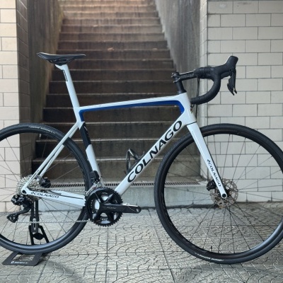 Bicicleta de estrada Colnago branca, azul e preta com guiador curvo preto em chão de azulejos