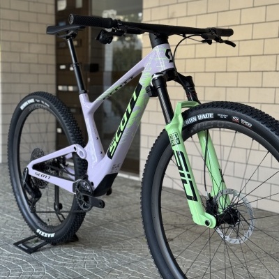 Bicicleta de montanha Scott em lilás e verde, com pneus Maxxis e suspensão SID, junto a parede de tijolos claros