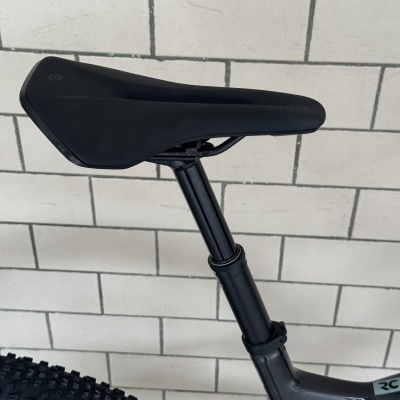 Selim de bicicleta preto com haste preta e quadro cinza escuro junto a parede de azulejo branco.