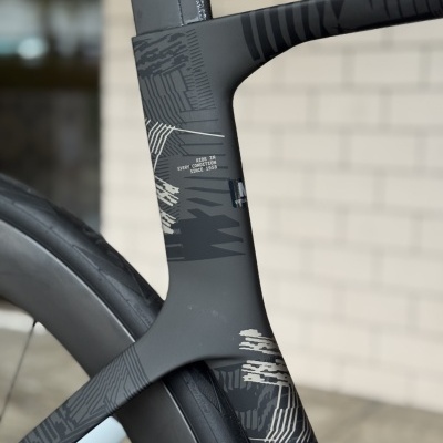 Quadro de bicicleta de carbono cinza escuro com padrão gráfico preto e cinza ao lado de roda preta, texto branco visível no quadro.