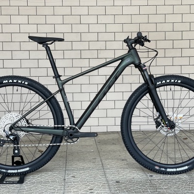 Bicicleta de montanha cinza escuro com pneus pretos Maxxis junto a parede de azulejos brancos