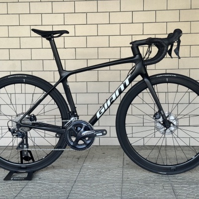 Bicicleta de estrada preta com quadro carbono, rodas CONTINENTAL, grupo Ultegra junto a parede de azulejos claros.
