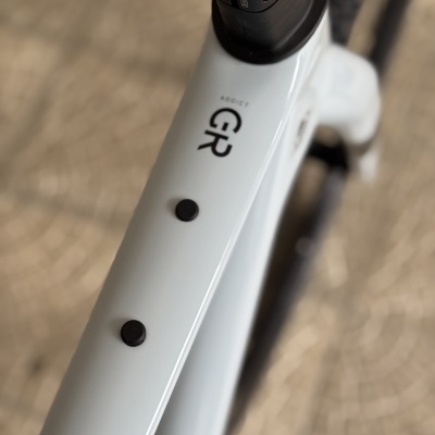 Quadro branco de bicicleta com texto 'AGGRESSIVE GR' e dois botões pretos, chão pavimentado desfocado ao fundo.