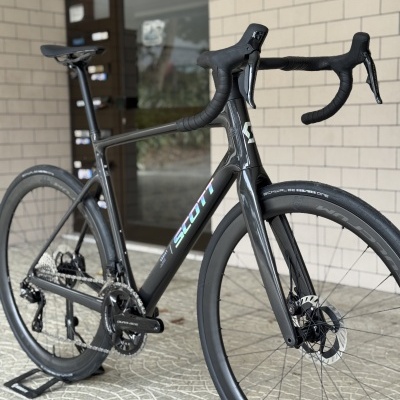 Bicicleta de estrada Scott preta com rodas de disco e guiador drop num piso de azulejos em frente a uma porta de vidro