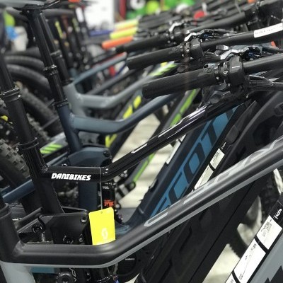 Várias bicicletas pretas e cinza alinhadas em loja com selins etiquetados e marcas visíveis.