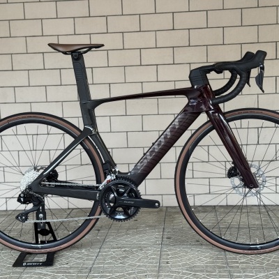 Bicicleta de estrada aerodinâmica preta e castanha com pneus castanhos, componentes Shimano 105, apoiada em suporte preto em frente a parede de azulejos brancos.
