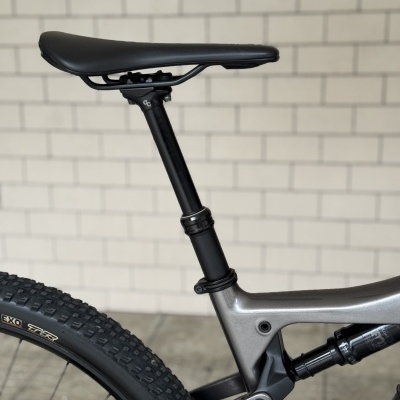 Assento de bicicleta preto, quadro cinza metálico, pneu traseiro preto com texto EXO TR, suspensão traseira, fundo de azulejos.