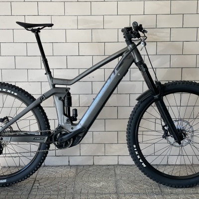 Bicicleta elétrica de montanha cinza escuro com pneus Maxxis junto a parede de azulejos brancos