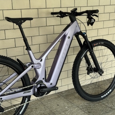 Bicicleta elétrica de montanha SCOTT lilás com pneus Maxxis em frente a parede de azulejos bege.