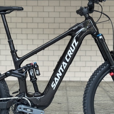 Bicicleta de montanha preta Santa Cruz com suspensão Fox e pneus Maxxis num ambiente com parede cinza.