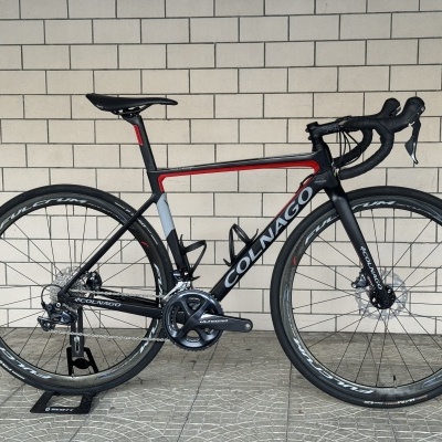 Bicicleta de estrada Colnago com quadro preto, vermelho e cinza diante de parede de azulejos brancos