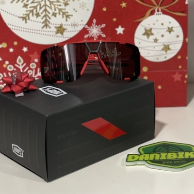 Óculos de ciclismo com lente escura e estrutura vermelha em caixa preta com logotipo, autocolante Danibikes, fundo decorativo natal.