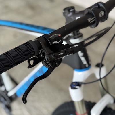 Guiador de bicicleta com punho preto, alavanca de travão Shimano e seletor SLX, quadro azul e branco ao fundo