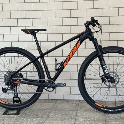 Bicicleta de montanha preta com detalhes laranja encostada a parede cinza