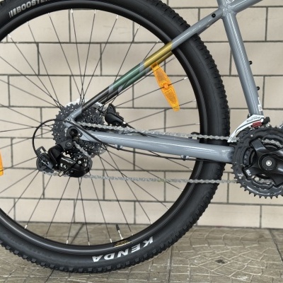 Roda traseira de bicicleta com pneu preto e quadro cinzento metálico junto a parede de azulejos brancos