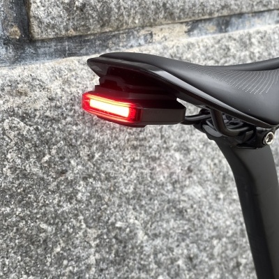 Luz traseira vermelha montada sob selim de bicicleta preto junto a parede de pedra