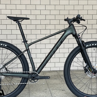 Bicicleta de montanha cinza escuro com pneus Maxxis pretos encostada a parede de azulejos