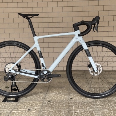 Bicicleta de estrada cinzenta com rodas pretas encostada a um suporte em calçada junto a parede de tijolos