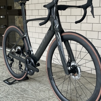 Bicicleta de estrada preta em carbono com pneus castanhos apoiada numa parede branca