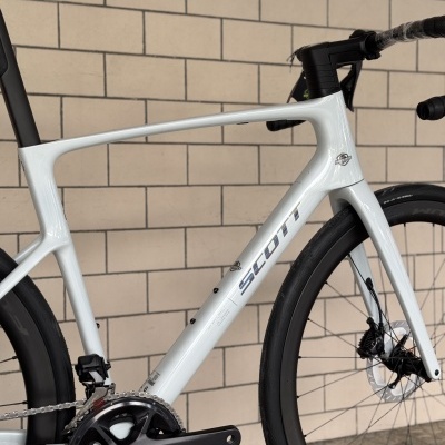 Bicicleta de estrada branca SCOTT com pedaleira ULTEGRA e pneus pretos junto a parede de azulejos bege