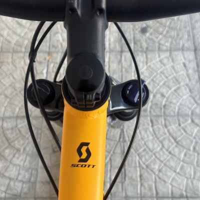 Bicicleta amarela SCOTT com guiador preto e suspensão dianteira, vista de cima sobre calçada cinza.