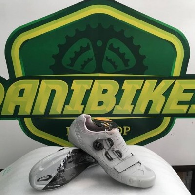 Sapatilhas de ciclismo brancas com roscas e velcro sobre suporte, cartaz DANI BIKES ao fundo