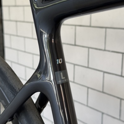 Quadro preto brilhante de bicicleta de estrada em carbono com etiquetas 10 e L VIRTUAL 58 CM contra parede de azulejos brancos