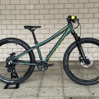 Bicicleta de montanha Scott verde escuro com detalhes amarelos ao lado de parede de azulejos.