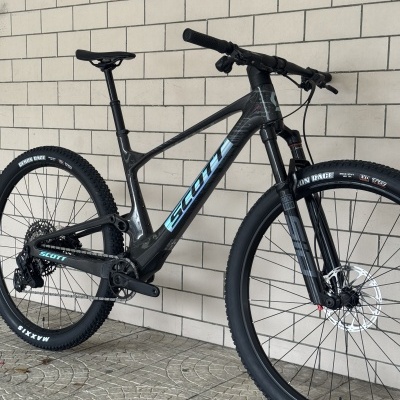 Bicicleta de montanha preta e azul claro SCOTT com pneus Maxxis e suspensão RockShox junto a uma parede de azulejos brancos.