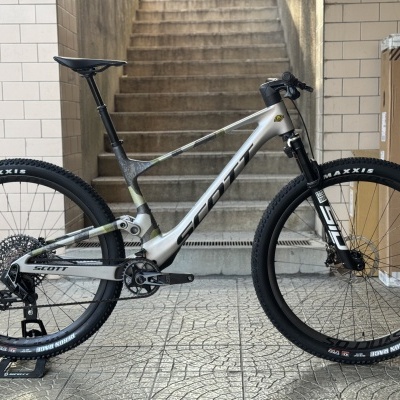 Bicicleta de montanha Scott prateada com pneus Maxxis negros em ambiente com escadas e azulejos.