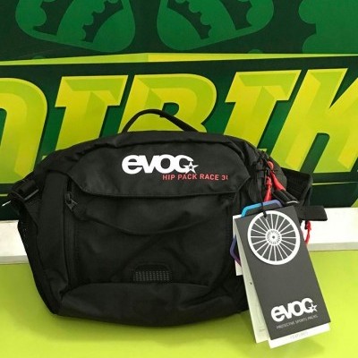Mala de cintura preta EVOC HIP PACK RACE 3L com etiquetas penduradas em fundo verde e amarelo