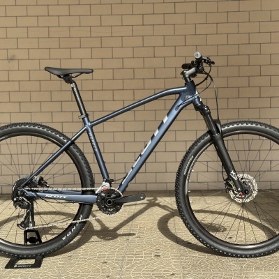 Bicicleta montanha azul metálico com pneus pretos e suspensão, apoiada num suporte preto