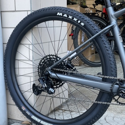 Roda traseira de bicicleta de montanha com pneu preto MAXXIS e quadro cinza fosco junto a parede de azulejos brancos