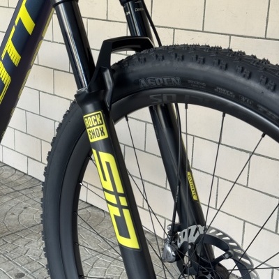 Parte frontal de bicicleta com suspensão Rock Shox preta e amarelo, pneu grossos com cravos, fundo parede de azulejos brancos.