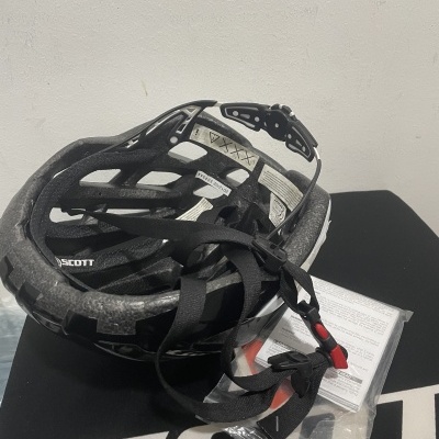 Capacete ciclismo preto e branco Scott com tiras ajustáveis e embalagem em superfície preta com logo