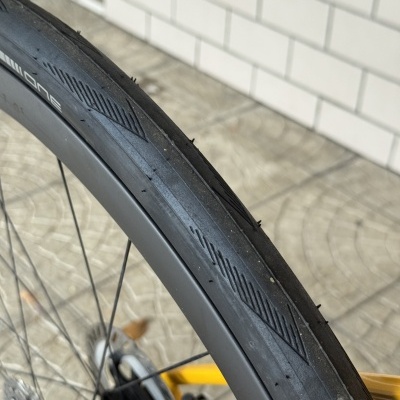 Pneu de bicicleta preta com aro cinza e raios metálicos em fundo de chão calcário e parede de azulejos brancos