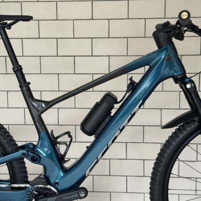 Bicicleta de montanha azul e preta com garrafa térmica e pneus Maxxis encostada a parede