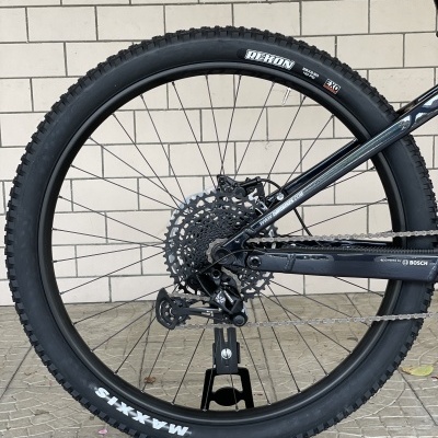 Roda traseira de bicicleta com pneu MAXXIS REKON 29x2.40 em suporte preto, com parede de tijolos ao fundo.