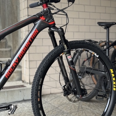 Bicicleta de montanha negra com pneus Maxxis e suspensão Rock Shox encostada a parede com azulejos.