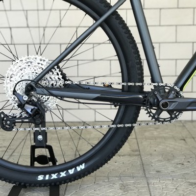 Parte traseira de uma bicicleta com pneu Maxxis preto, corrente e cassete metálicos, quadro cinza escuro em piso cinzento