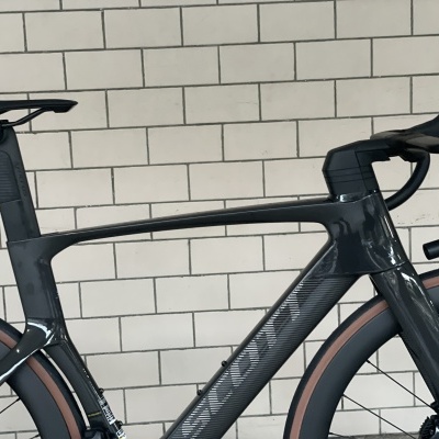 Bicicleta de estrada preta com detalhes castanhos junto a uma parede de tijolos brancos