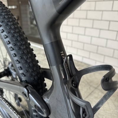 Quadro cinza escuro de bicicleta de montanha com roda traseira e suporte de bidon preto em fundo de parede de azulejos brancos.