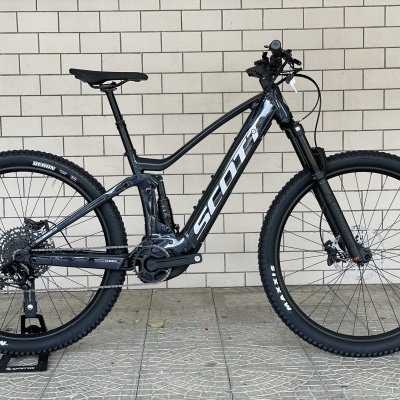 Bicicleta de montanha eléctrica SCOTT cinza escuro com pneus Maxxis e travões de disco junto a uma parede de tijolos.