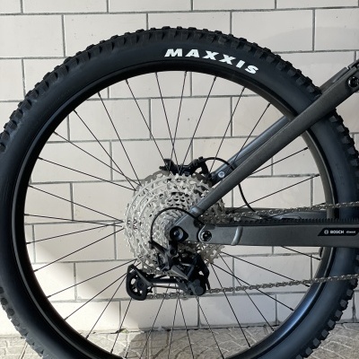 Roda traseira de bicicleta com pneu Maxxis preto e quadro cinza junto a parede branca