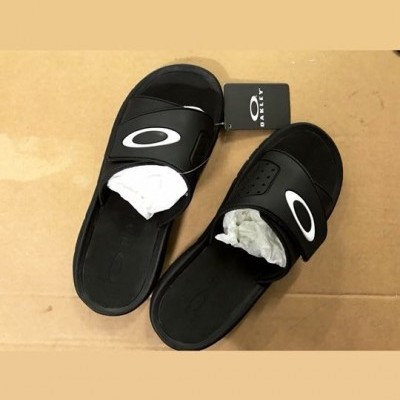 Par de chinelos pretos com logo branco e etiqueta Oakley sobre superfície castanha