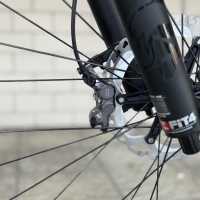 Roda de bicicleta com garfo preto, disco metálico de travão e pinça com texto, em chão de pedra